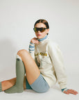 Person wearing sunglasses, a white sweater, and green knee-high socks on a plain background