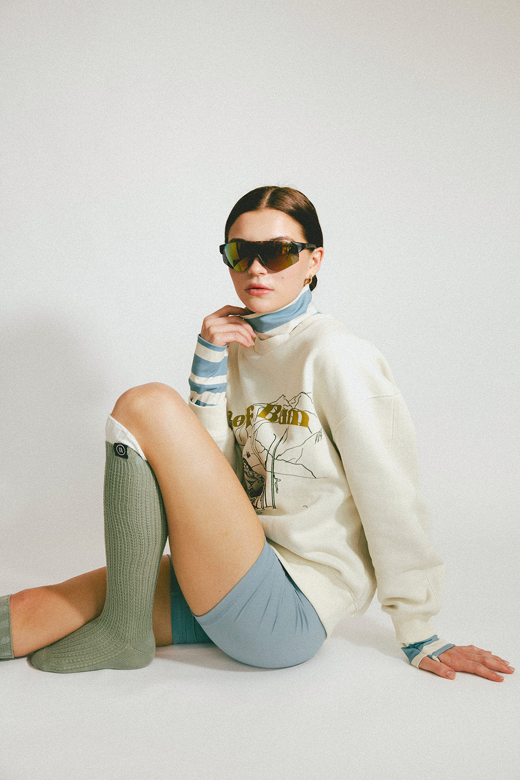 Person wearing sunglasses, a white sweater, and green knee-high socks on a plain background