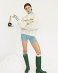 Person wearing a cream sweatshirt with text, light blue shorts, and green rain boots on a white background