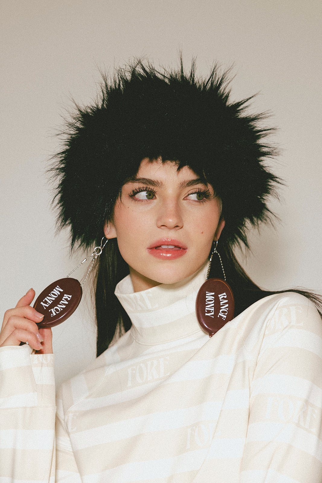 Woman wearing a black furry hat and holding brown circular objects with text, against a plain background.