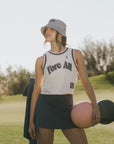 Woman in a white tank top with 'Fore All' text, indigo skirt, and patterned hat standing on a golf course.