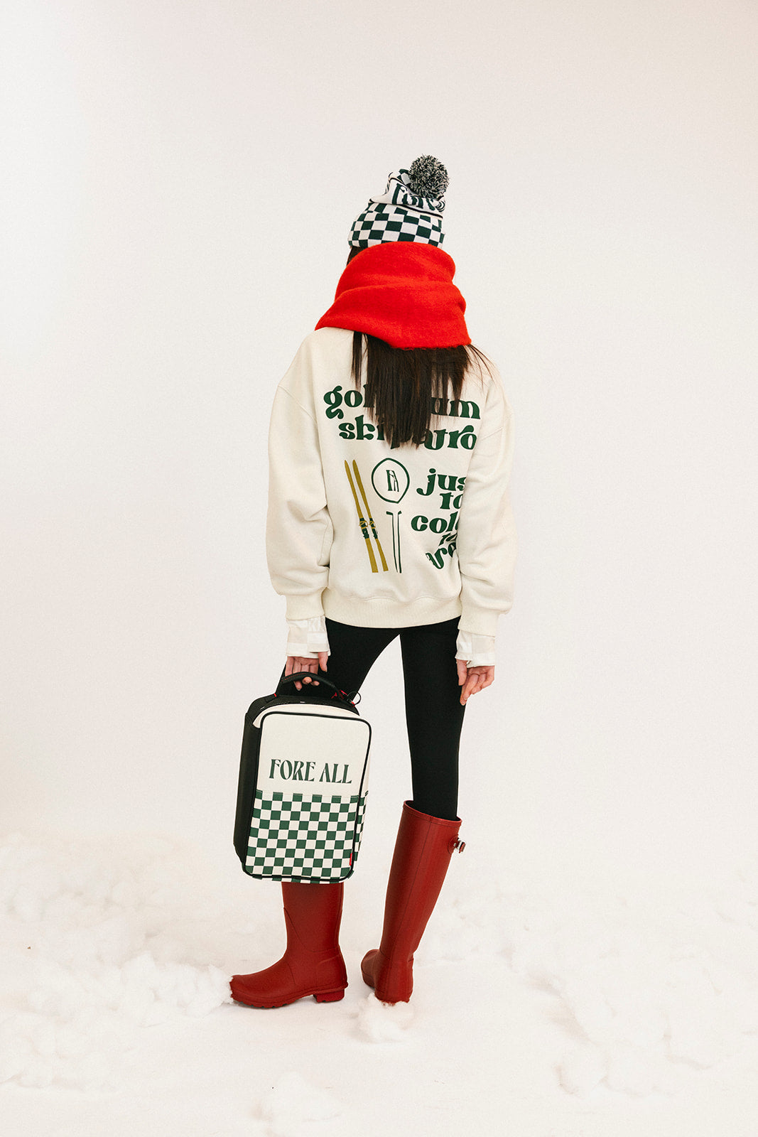 Person wearing a white sweatshirt with text, black leggings, red rain boots, and a checkered hat, holding a bag with a white background.