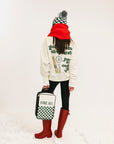 Person wearing a white sweatshirt with text, black leggings, red rain boots, and a checkered hat, holding a bag with a white background.
