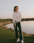 Woman in a checkered sweater and green pants holding a golf club on a golf course.