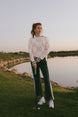 Woman in a checkered sweater and green pants holding a golf club on a golf course.