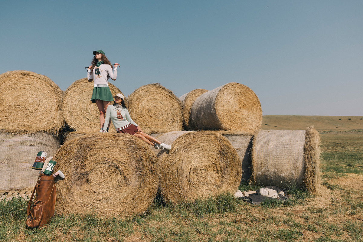 Two girls posing outdoors with large hay bales under a clear blue sky. One girl stands on a hay bale wearing a white long-sleeve top, green skirt, and black cap, while the other sits on a hay bale wearing a light green top and red skirt.