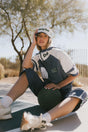 Woman sitting on a court wearing an indigo and white outfit with a cap, outdoors.