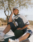 Woman sitting on a court wearing an indigo and white outfit with a cap, outdoors.