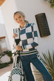 Woman holding a golf bag with an indigo checkered Dorothy sweater with a checkered pattern, standing outdoors.