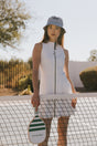 Woman holding a paddle on a tennis court wearing a white dress and patterned hat.
