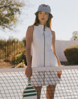 Woman holding a paddle on a tennis court wearing a white dress and patterned hat.