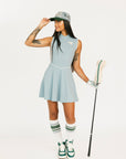 Woman in light blue sleeveless dress and green sneakers holding a golf club on a white background