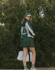A woman wearing a long-sleeved green and pink warm-up jacket with a hood, zipper pockets, and colorblocking. She also sports a matching green cap.