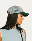 Woman wearing a blue and green cap with a logo, against a plain background