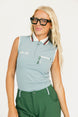 Woman wearing a sleeveless light blue golf polo shirt with a logo and green pants on a white background