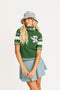 Woman wearing a green Macbeth sweater with a Fore All logo and a light blue skirt on a white background