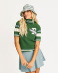 Woman wearing a green Macbeth sweater with a Fore All logo and a light blue skirt on a white background