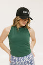 A woman wearing a green tank top with a built-in bra, paired with patterned shorts and a black cap with the text 'FORE ALL' on it.