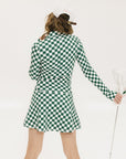 A person wearing a green and white checkered mock-neck long-sleeve golf top and matching golf skort, holding a golf club.