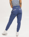 A female model wearing a sleeveless top with a geometric pattern and blue athletic pants, standing against a white background.