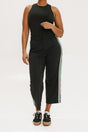 A female model wearing black sleeveless top and black crop pants with white side stripes, paired with black shoes.