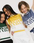 Three models wearing a sleeveless mock-neck sweater with intarsia knit design, featuring horizontal green, mustard, and indigo stripes and the word 'FORE' printed across the chest.