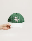 A green baseball cap with pinstripe detailing and an embroidered logo held in a hand against a white background.