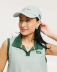 A woman wearing a green seersucker baseball cap with 'Chasing Balls' text embroidered on it and an oversized bow at the back.