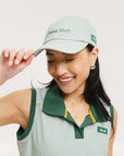 A woman wearing a green seersucker baseball cap with 'Chasing Balls' text embroidered on it and an oversized bow at the back.