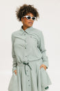 A woman wearing a green striped button-up shirt with a collar, sleeve cuffs, and side seam hem, paired with a matching skirt, sunglasses, and white shoes.