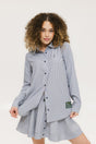 A woman wearing a long, button-up shirt with indigo stripes, featuring a collar, button-down front, and elastic toggles at the side seam hem and sleeve cuffs.