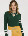 A woman wearing a green ribbed keyhole polo with white stripe accents on the sleeves and a gold smiley face button at the center front neck, paired with a yellow and white skirt.