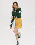 A woman wearing a green ribbed keyhole polo with white stripe accents on the sleeves and a gold smiley face button at the center front neck, paired with a yellow and white skirt.