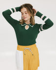 A woman wearing a green ribbed keyhole polo with white stripe accents on the sleeves and a gold smiley face button at the center front neck, paired with a yellow and white skirt.