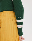 A woman wearing a green ribbed keyhole polo with white stripe accents on the sleeves and a gold smiley face button at the center front neck, paired with a yellow and white skirt.