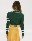 A woman wearing a green ribbed keyhole polo with white stripe accents on the sleeves and a gold smiley face button at the center front neck, paired with a yellow and white skirt.