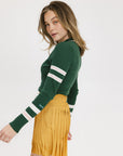 A woman wearing a green ribbed keyhole polo with white stripe accents on the sleeves and a gold smiley face button at the center front neck, paired with a yellow and white skirt.