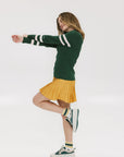A woman wearing a green ribbed keyhole polo with white stripe accents on the sleeves and a gold smiley face button at the center front neck, paired with a yellow and white skirt.