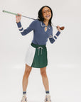 A woman wearing a blue ribbed keyhole polo with white stripe accents on the sleeves and a green and white pleated skirt.