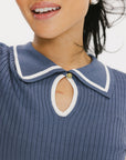 A woman wearing a blue ribbed keyhole polo with white stripe accents on the sleeves and a green and white pleated skirt.