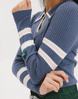 A woman wearing a blue ribbed keyhole polo with white stripe accents on the sleeves and a green and white pleated skirt.
