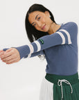 A woman wearing a blue ribbed keyhole polo with white stripe accents on the sleeves and a green and white pleated skirt.