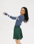 A woman wearing a blue ribbed keyhole polo with white stripe accents on the sleeves and a green and white pleated skirt.