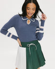 A woman wearing a blue ribbed keyhole polo with white stripe accents on the sleeves and a green and white pleated skirt.