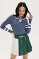 A woman wearing a blue ribbed keyhole polo with white stripe accents on the sleeves and a green and white pleated skirt.