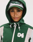 A woman wearing a long-sleeved green and pink warm-up jacket with a hood, zipper pockets, and colorblocking. She also sports a matching green cap and appears to be standing against a white background.