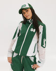 A woman wearing a long-sleeved green and pink warm-up jacket with a hood, zipper pockets, and colorblocking. She also sports a matching green cap and appears to be standing against a white background.