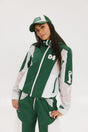 A woman wearing a long-sleeved green and pink warm-up jacket with a hood, zipper pockets, and colorblocking. She also sports a matching green cap and appears to be standing against a white background.