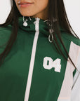 A woman wearing a long-sleeved green and pink warm-up jacket with a hood, zipper pockets, and colorblocking. She also sports a matching green cap and appears to be standing against a white background.