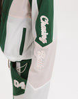 A woman wearing a long-sleeved green and pink warm-up jacket with a hood, zipper pockets, and colorblocking. She also sports a matching green cap and appears to be standing against a white background.